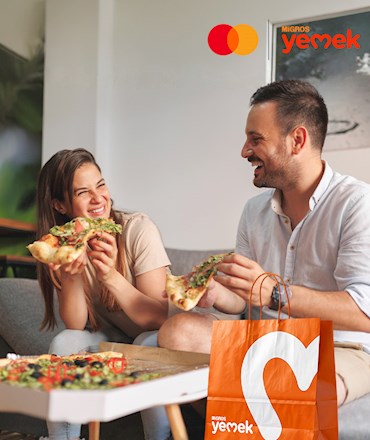 MİGROS yemek ve Mastercard logosu altında, evde kutudan pizza yiyerek neşeyle gülen bir kadın ve erkek ile önlerinde Migros Yemek poşeti.