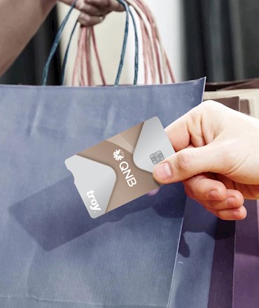 troy QNB logolu bir kredi kartını tutan el ve arka planda alışveriş çantaları taşıyan biri.