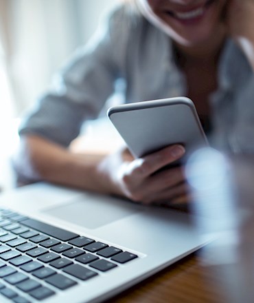 Gülümseyen bir kişi dizüstü bilgisayarın önünde akıllı telefon kullanıyor.