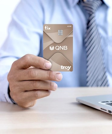 fix KOBİ QNB troy. Gömlek ve kravatlı bir kişi, bilgisayar başında elinde bronz renkli kredi kartını tutuyor.